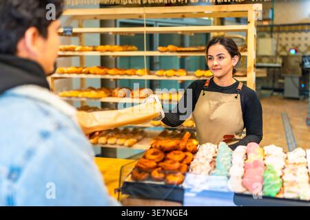 Hispanic female baker in apron handing bread to customer while standing at counter in bakery Stock Photo