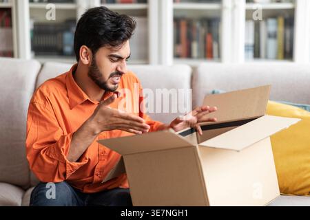Disappointed Middle Eastern man gesturing hands opening parcel sitting on couch at home Stock Photo