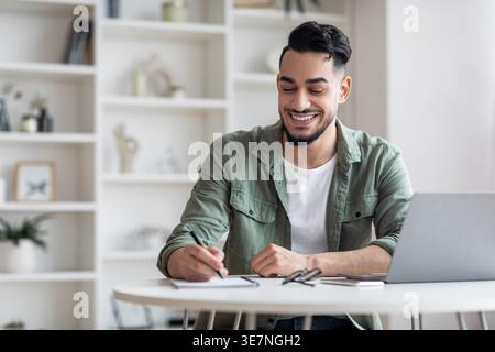 Smiling Arab man writing down notes sitting at table with laptop at home Stock Photo
