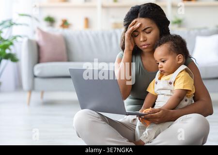Black mother with baby boy touching forehead suffering from headache holding laptop sitting with crossed legs on floor in living room at home Stock Photo