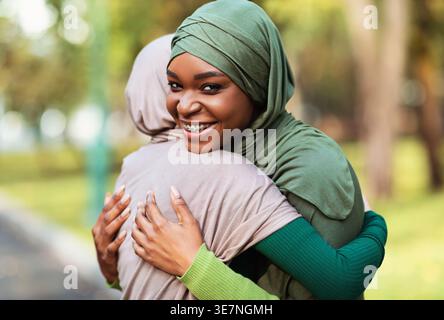 Smiling Black and Middle Eastern women wearing hijabs embracing each other while standing in city park during daytime Stock Photo