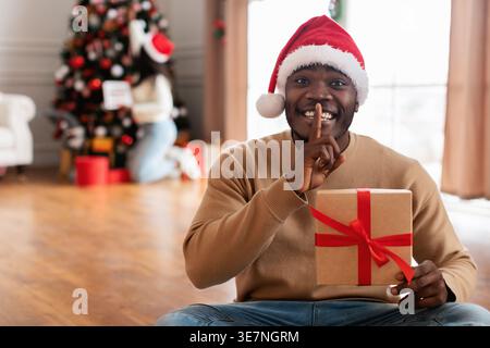 Black man wearing Santa hat holding gift gesturing silence with finger on lips sitting on floor near wife decorating Chirstmas tree in living room ... Stock Photo