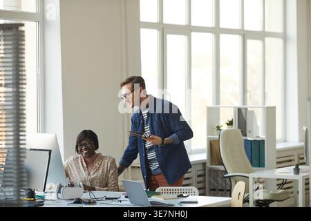Black female programmer wearing eyeglasses working on computer with caucasian male colleague talking to her while sitting with virtual reality head... Stock Photo