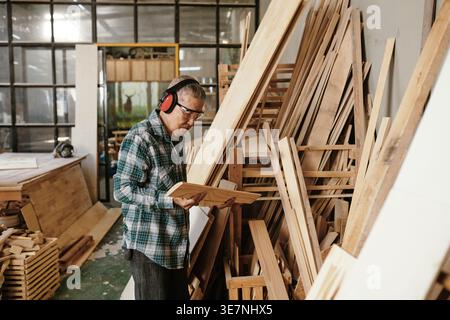 Senior Asian male carpenter in protective googles and earmuffs looking and holding wooden piece while standing sideways on workshop Stock Photo