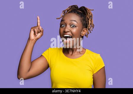 Black woman with locks wearing yellow shirt pointing finger up having and idea while standing against purple background in studio Stock Photo