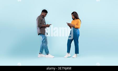 Black man and woman using smartphones standing against blue background in studio Stock Photo