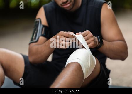 Black man wearing sportswear with armband smartphone wrapping injured knee while sitting outdoors during daytime Stock Photo