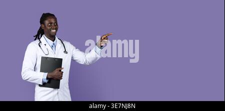 Smiling Black male doctor with dreads wearing white suit with stethoscope holding clipboard pointing away while standing against purple background ... Stock Photo