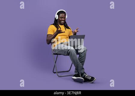 Cheerful stylish long-haired young black man sitting on chair, using brand new computer and wireless headset, having video chat with colleagues or ... Stock Photo