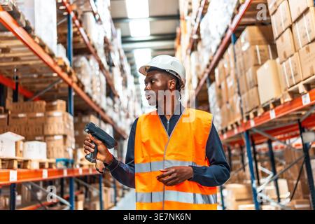 Black male worker with dreads wearing safety vest and hard hat scanning package codes using wireless scanner while standing in logistic warehouse Stock Photo