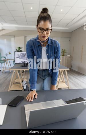 Female in late 20s leaning over laptop in design studio, reviewing plans on monitor with notebook Stock Photo