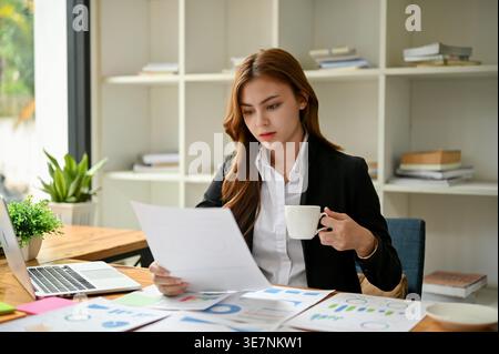 Serious Asian business woman in black suit looking at reports and having cup of coffee while sitting at table in office Stock Photo