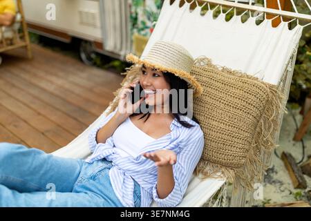 Cheerful Asian woman wearing hat having phone call looking away gesturing hand while lying on hammock near motorhome during camping trip in forest Stock Photo