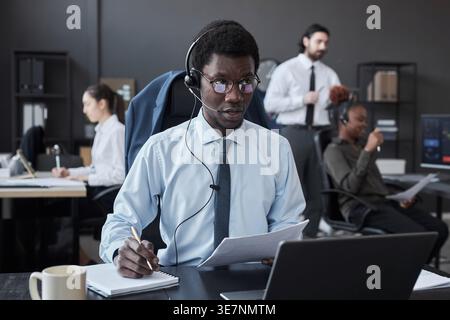 Black male broker wearing eyeglasses and headset talking to customer and writing notes while sitting at his desk with laptop in office Stock Photo