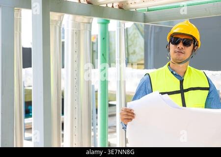 Asian male engineer wearing safety helmet looking up holding blueprints standing near pipeline on rooftop during daytime Stock Photo