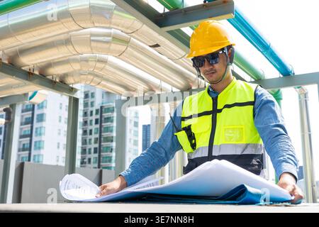 Asian male engineer wearing safety helmet checking blueprints standing at table near pipeline on rooftop during daytime Stock Photo