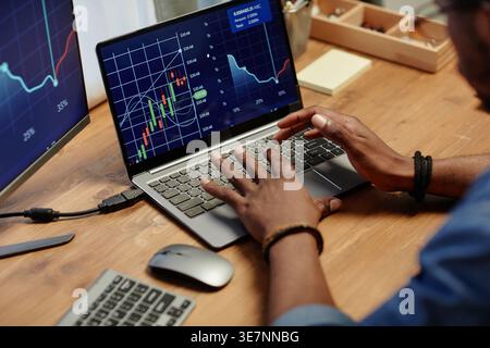 Cropped image of Black male broker typing on laptop keyboard and analyzing financial graph on screen while sitting at wooden table in office Stock Photo