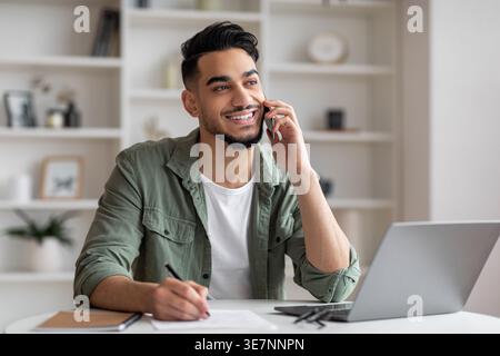 Arab man looking away having phone call writing down notes sitting at table with laptop at home Stock Photo