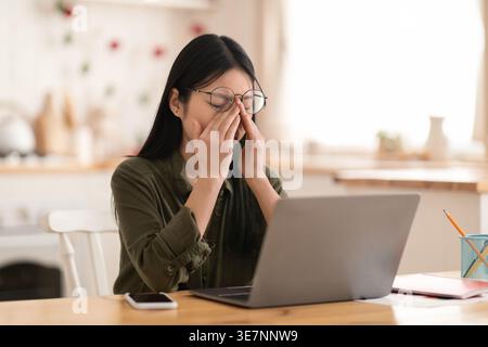 Tired overworked young korean woman wearing eyeglasses digital nomad rubbing eyes, using laptop at kitchen, asian lady working from home, have deadlin Stock Photo
