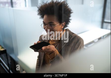 Smiling Black businessman with afro wearing eyeglasses recording voice message using smartphone standing in office Stock Photo