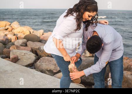 Hispanic son leaning forward helping his mother wearing sandals while standing near beach at pier during daytime Stock Photo