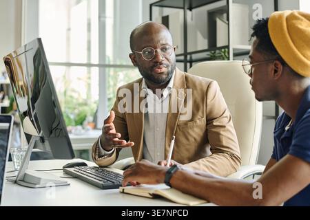 Serious Black male programmers discussing new project while sitting at desk with computer in office Stock Photo
