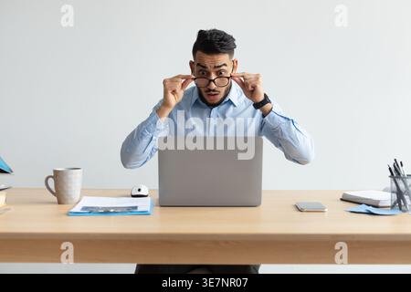Error. Portrait of shocked scared Arab guy sitting at desk using laptop, taking off glasses looking at pc screen in amazement. Stressed man suffering Stock Photo