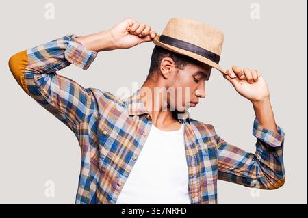 Black man with closed eyes tipping straw hat while standing against light gray background Stock Photo