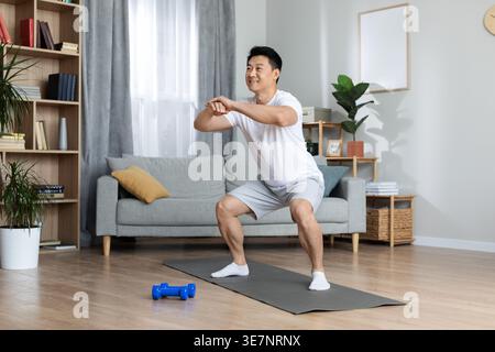 Asian man in white t-shirt doing squats in living room at home Stock Photo