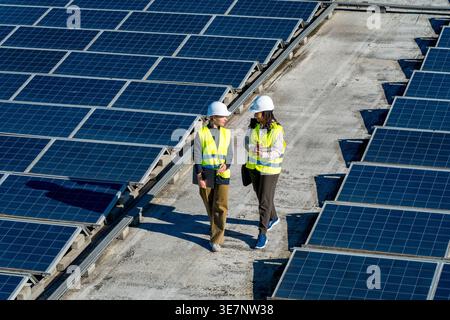 Hispanic and caucasian female engineers wearing hard hats and safety vests discussing work while walking on commercial rooftop with solar panels on... Stock Photo