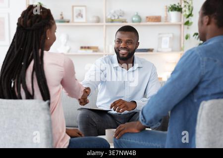 Black male psychologist sitting and shaking hands with Black couple after successful therapy session in office Stock Photo