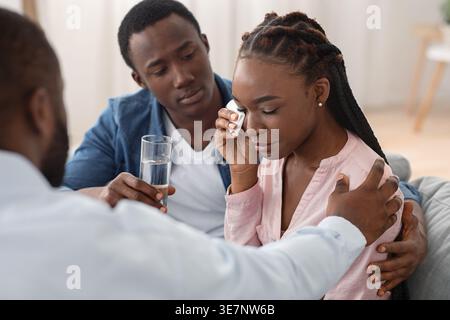 Black man giving water and comforting his wife having mental health problems while sitting on couch and talking with male therapist in office Stock Photo