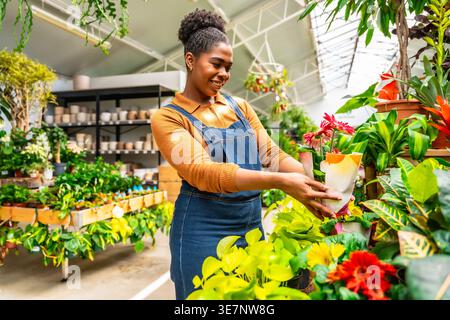 Hispanic female gardener in denim overalls holding potted plant and smiling while standing in greenhouse Stock Photo
