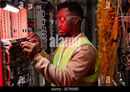 Black male network technician inspecting servers in data center lit by red neon light Stock Photo