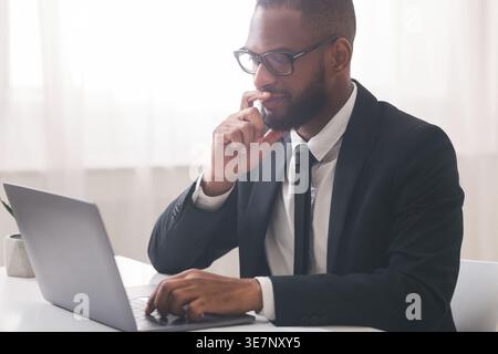 Black businessman wearing eyeglasses touching his chin and looking at laptop screen thinking while sitting at desk in office Stock Photo