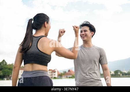 Cheerful Thai couple with raised arms and closed fists celebrating success after reaching their running target while standing together near lake in... Stock Photo