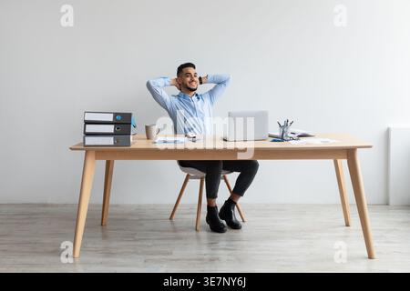 Relaxed Middle Eastern man with closed eyes and hands behind head sitting at table with laptop in office Stock Photo