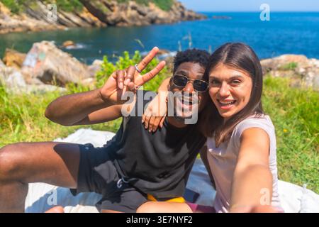 Black boyfriend with caucasian girlfriend taking selfie while having picnic sitting on blanket near seaside on sunny day Stock Photo