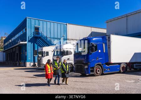 Diverse group of engineers wearing hard hats and safety vests walking together near transport trucks at cold storage warehouse on sunny day Stock Photo