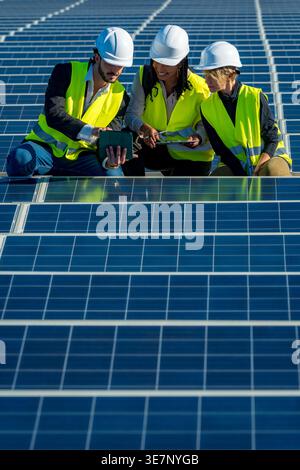 Diverse group of engineers wearing hard hats and safety vests discussing data on tablet while crouching and working on large solar panel installati... Stock Photo