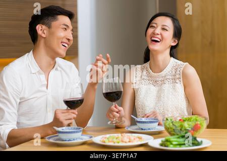 Cheerful Chinese couple holding glass of wine having a dinner while sitting at table at home Stock Photo