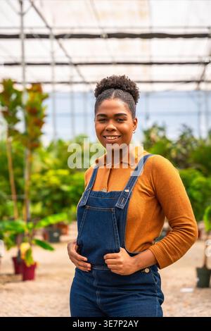 Hispanic female gardener in denim overalls smiling while standing sideways in greenhouse during daytime Stock Photo