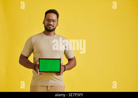 Asian man wearing eyeglasses showing tablet with green screen standing against yellow background in studio Stock Photo