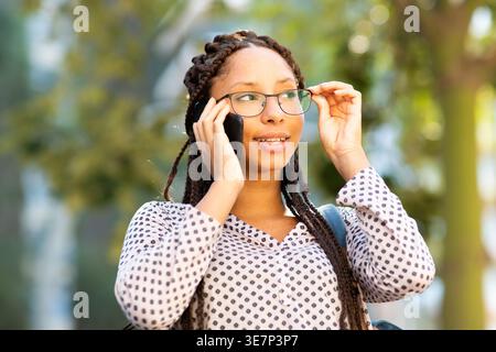 Smiling Mixed race woman with braids looking away having phone call while walking on street Stock Photo