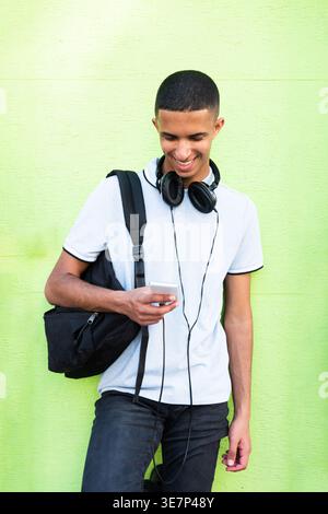 Smiling Arab male using cellphone leaning against green wall during daytime Stock Photo