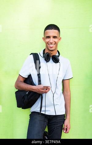 Smiling Arab male holding cellphone leaning against green wall during daytime Stock Photo