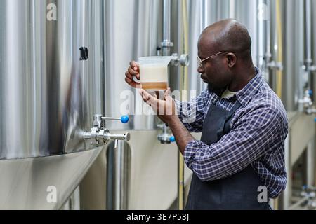 Focused Black bald man in apron examining pitcher with beer standing in factory Stock Photo