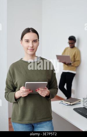 Smiling Mixed race woman holding tablet standing near table against Black man colleague leaning on wall using laptop in office Stock Photo