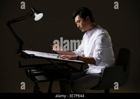 Chinese male architect working on construction plan sitting at table in office Stock Photo