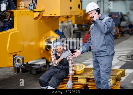 Hispanic female technician sitting on wooden platform injured with caucasian male technician having phone call in industrial factory during daytime Stock Photo